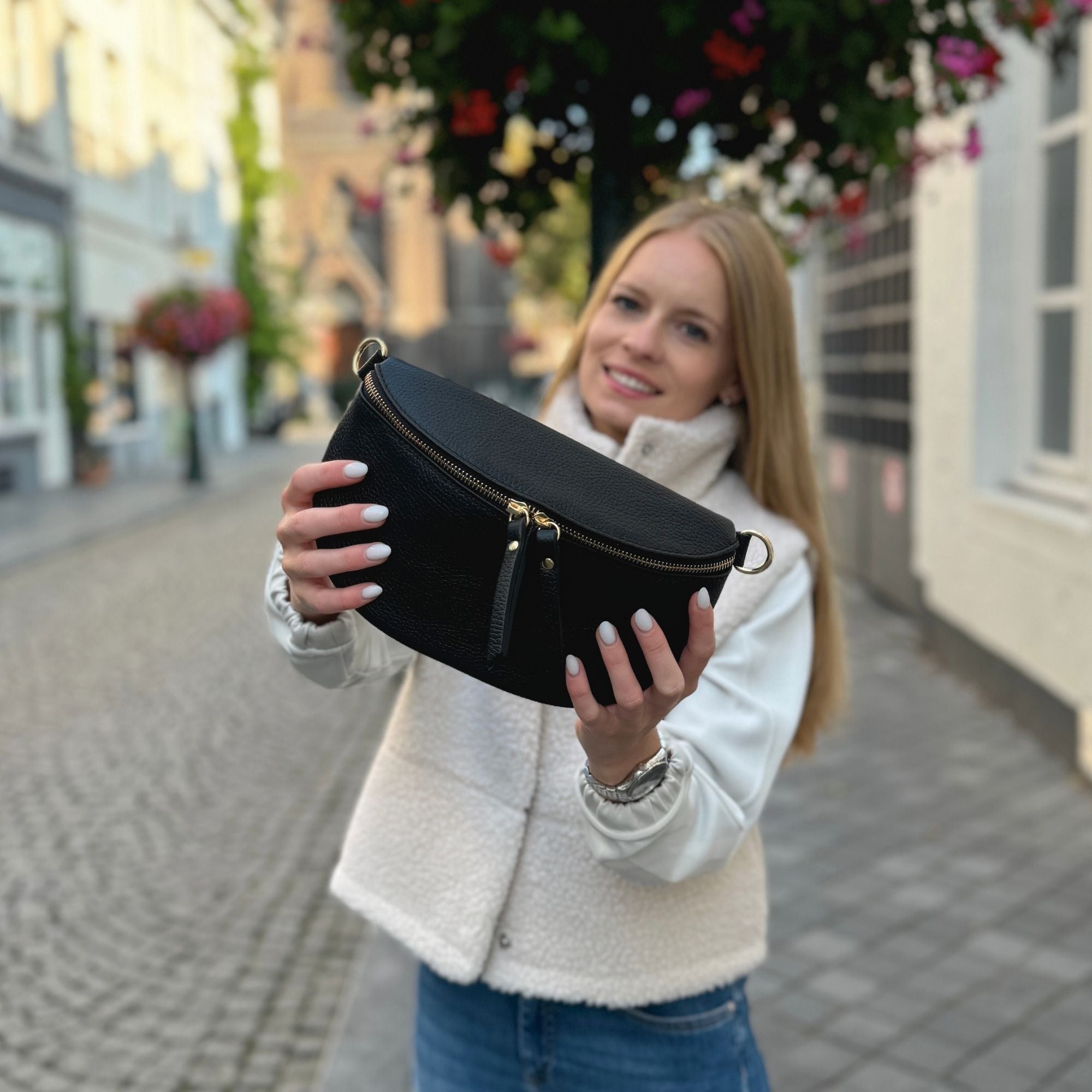 Schwarze Tasche mit goldfarbenem Verschluss im Vordergrund, Frau lacht im Hintergrund – fokussiertes Bild der Bauchtasche, gehalten mit einem Lächeln.