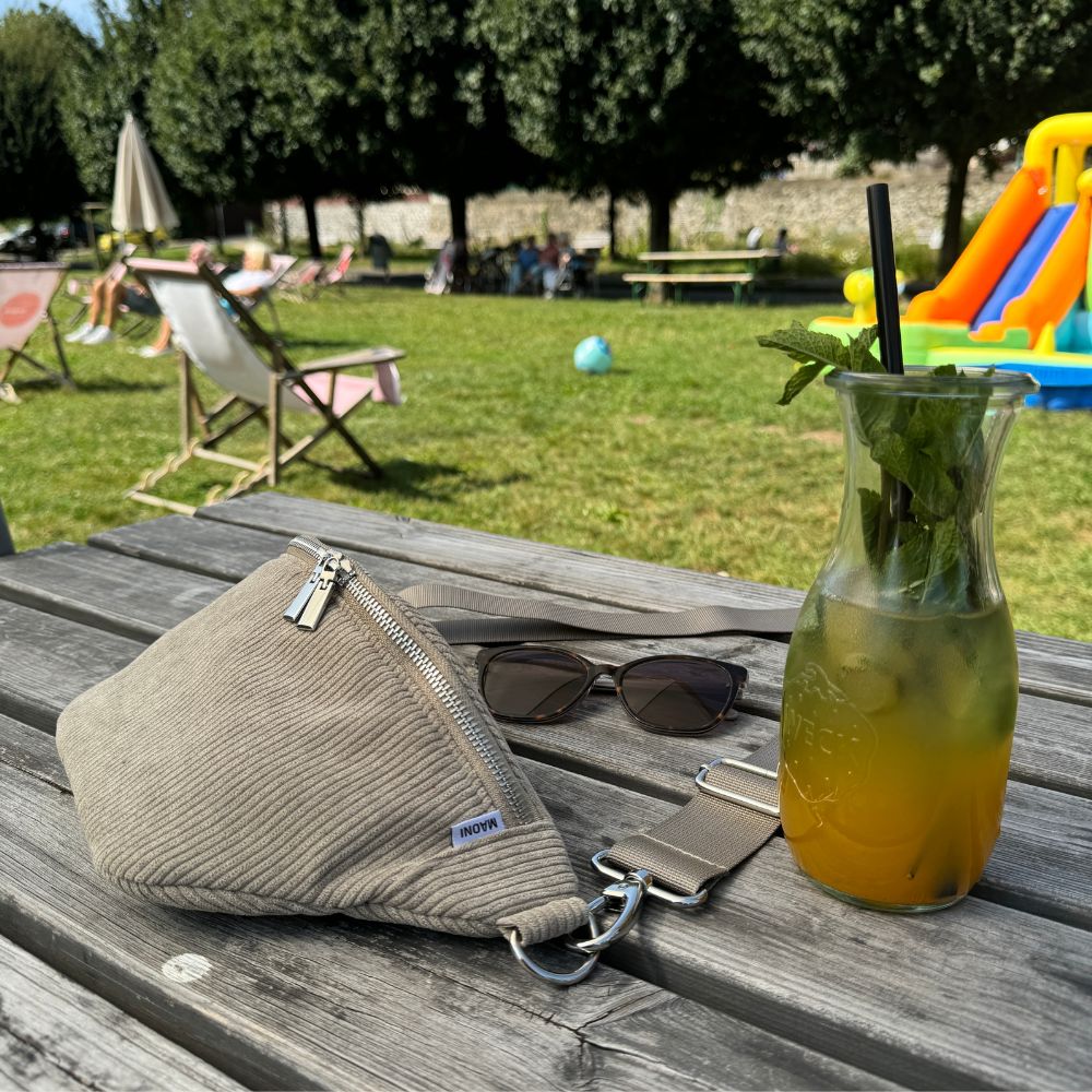 Entspannter Sommermoment: Cocktail, Sonnenbrille und Cord Bauchtasche auf einem Tisch, mit Sonnenliegen im Hintergrund.