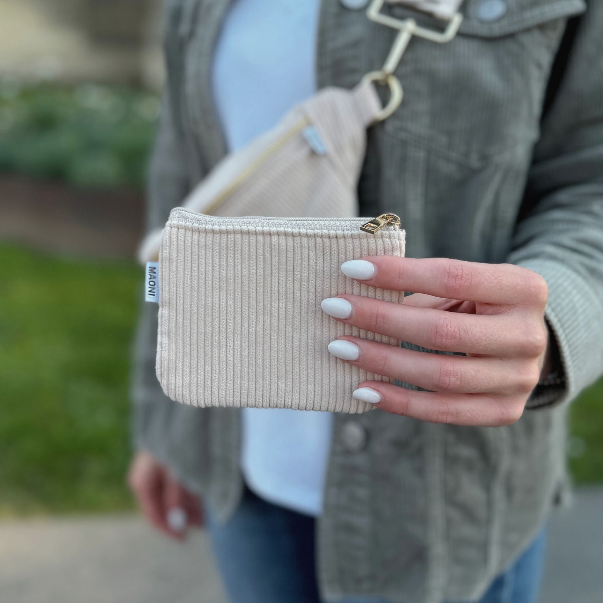 Frau hält Cord Etui in der Farbe Beige in die Kamera und trägt im Hintergrund die passende bauchtasche aus Cord und ein grünes cord Hemd.