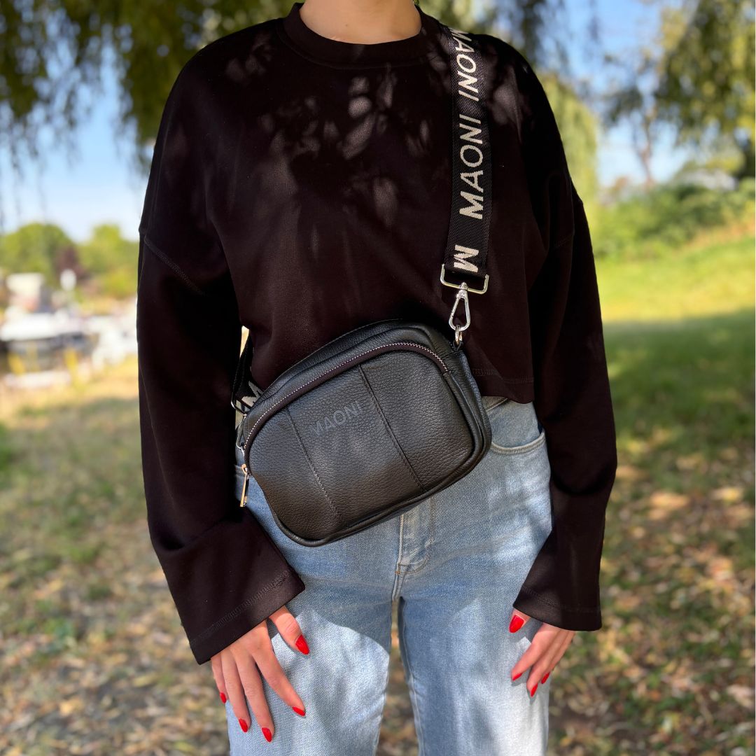 Frau trägt stylische Crossbody-Tasche in der Farbe schwarz über der Schulter, unterwegs im Grünen mit schwarzem Pullover und blauer Jeans