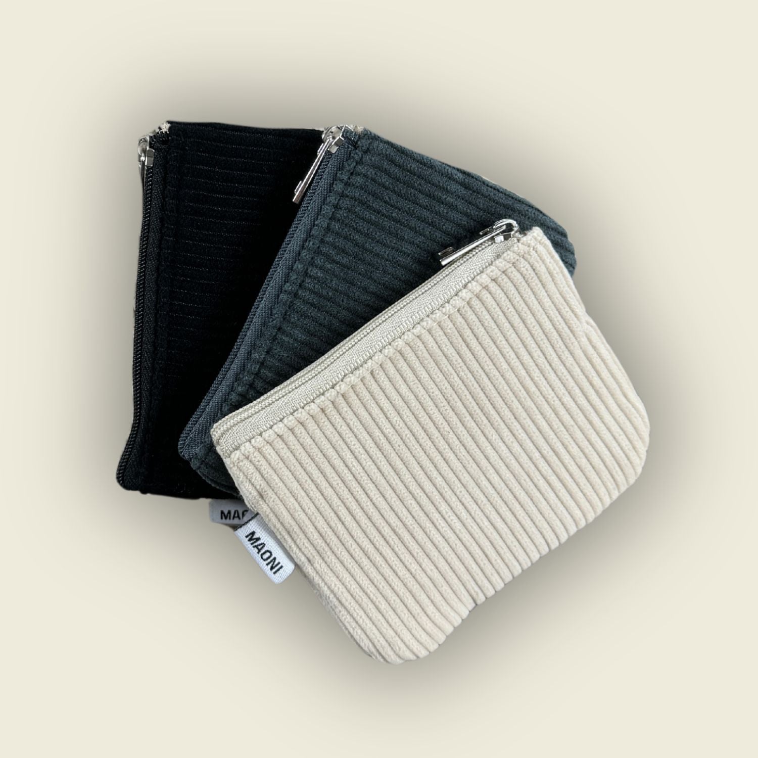 Einer Auswahl an Cord Etuis in dunkelgrau, beige und schwarz – modernes Accessoire ausgeschnitten auf beige Hintergrund.