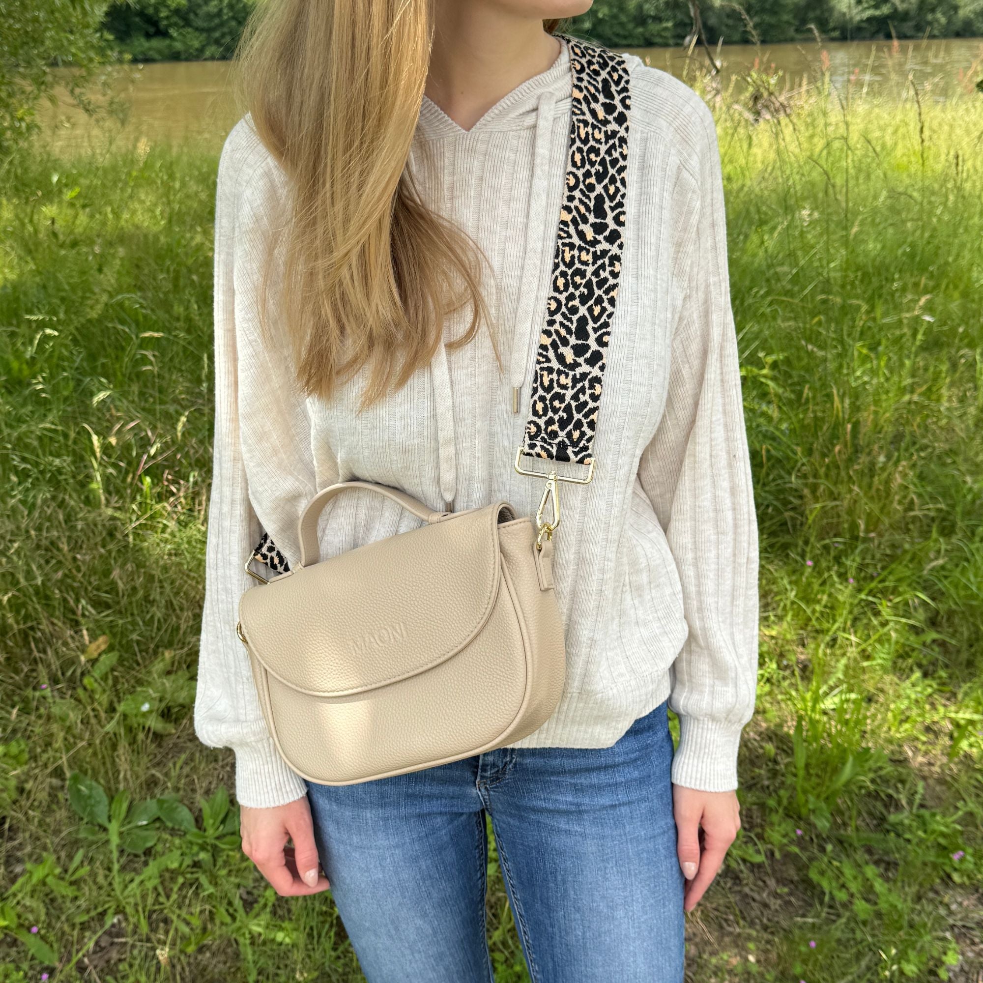 Frau mit blonden Haaren trägt taupefarbene Halfmoon-Tasche mit Leo-Gurt, kombiniert mit einem beigen Pullover und blauen Jeans.