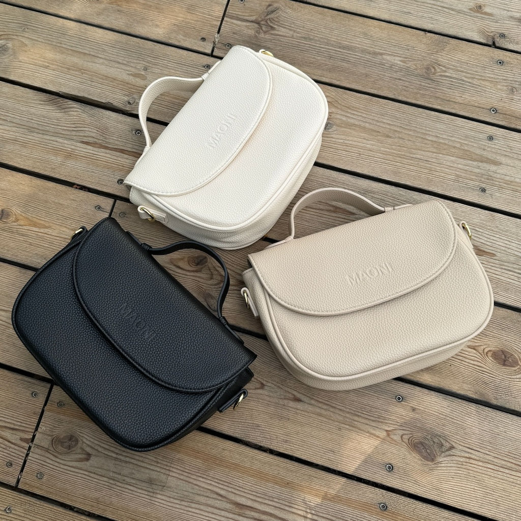 Drei Halfmoon Taschen liegen nebeneinander auf Holzoberfläche. Die Farben der Tasche sind beige, taupe und schwarz.