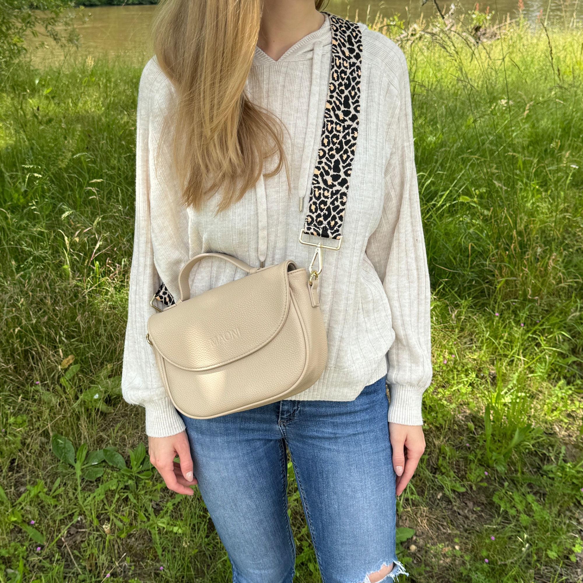 Frau trägt schwarze Halfmoon-Tasche in der Natur, kombiniert mit Jeans und beigem Pullover, dazu ein Leo-Gurt für den stylishen Look.