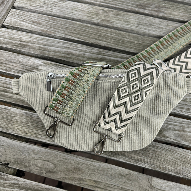 Grüne Cord-Bauchtasche liegt auf der Rückseite mit sichtbarem Reißverschlussfach auf der Rückseite für mehr Stauraum und Sicherheit. Auf der Tasche liegen zwei farblich passende Gurte in den Farben beige, dunkelgrün und braun.