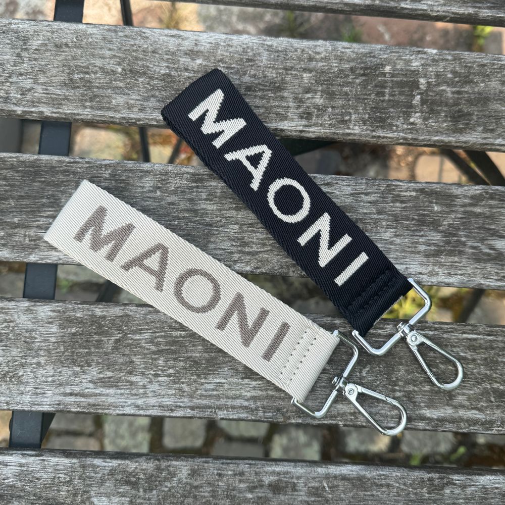 Beide MAONI-Schlüsselanhänger in Schwarz-Beige und Dunkeltaupe-Beige mit Logo-Schriftzug – dekorativ auf Holztisch arrangiert