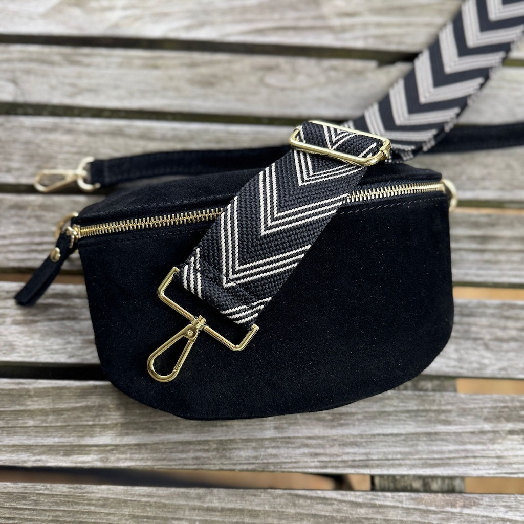 Schwarze Veloursleder Bauchtasche liegt auf Holztisch, darüber ein breiter gemusterter Gurt mit goldenem Verschluss