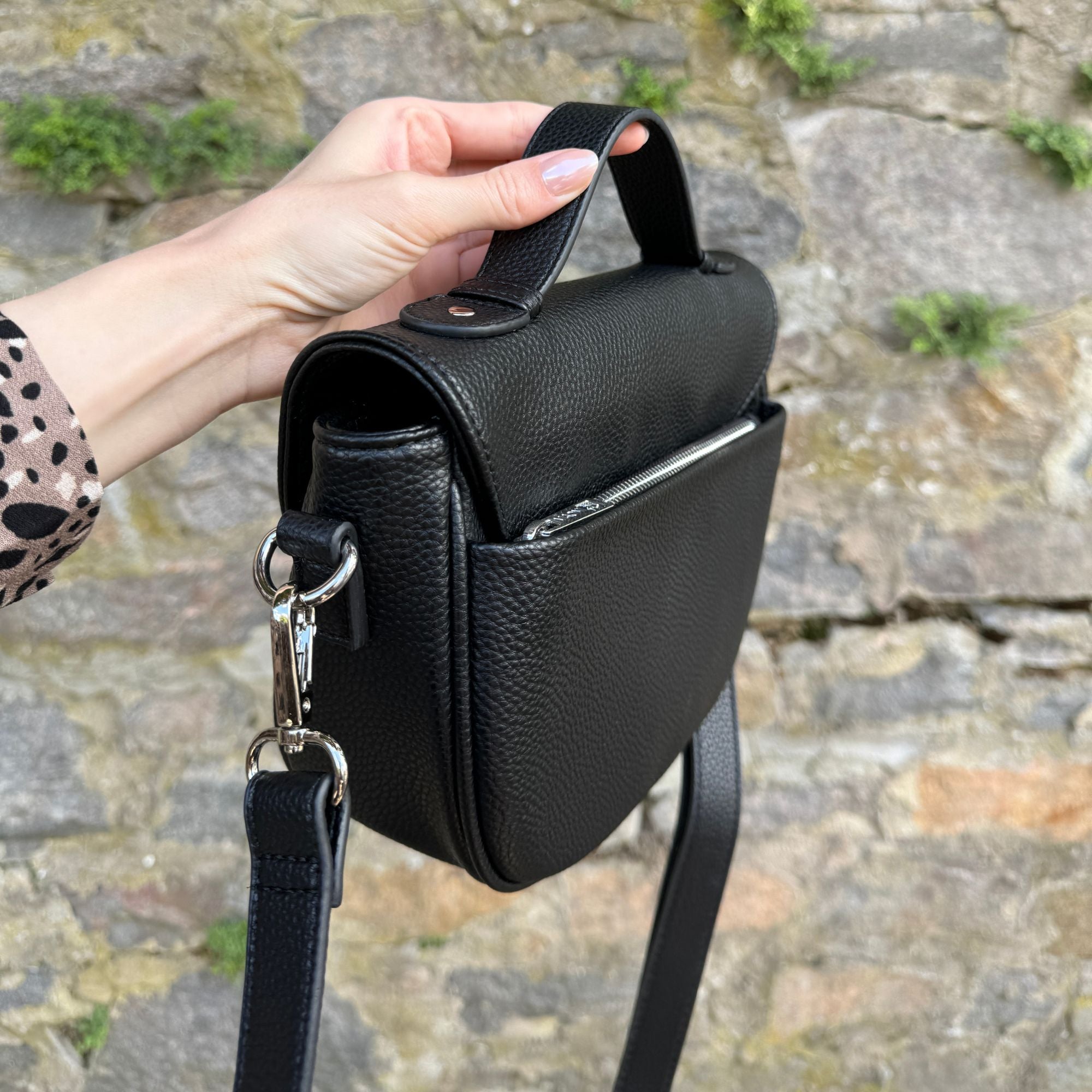 Schwarze Halfmoon-Tasche wird von Frau an kurzem Henkel gehalten, vor einer Steinwand, nur die Hand der Frau sichtbar.
