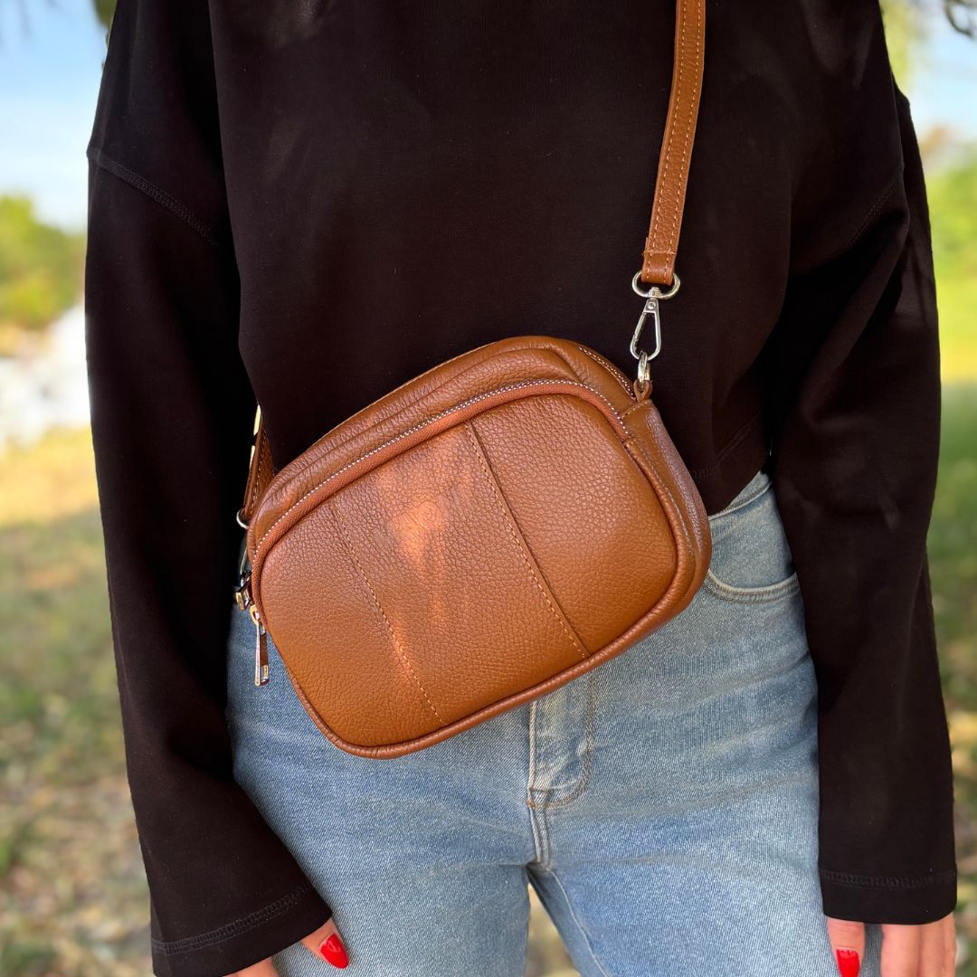 braune Crossbody-Tasche aus Leder, lässig getragen von einer Frau mit Jeans und schwarzem Pullover bei einem Spaziergang in der Natur