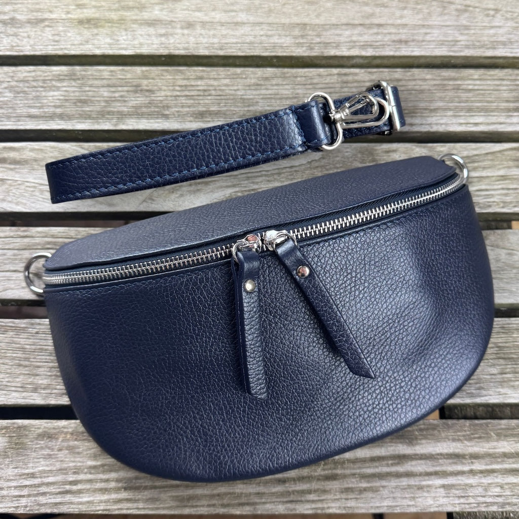 Große dunkelblaue Bauchtasche, von vorn liegend auf Holztisch – schmaler Ledergurt locker darübergelegt