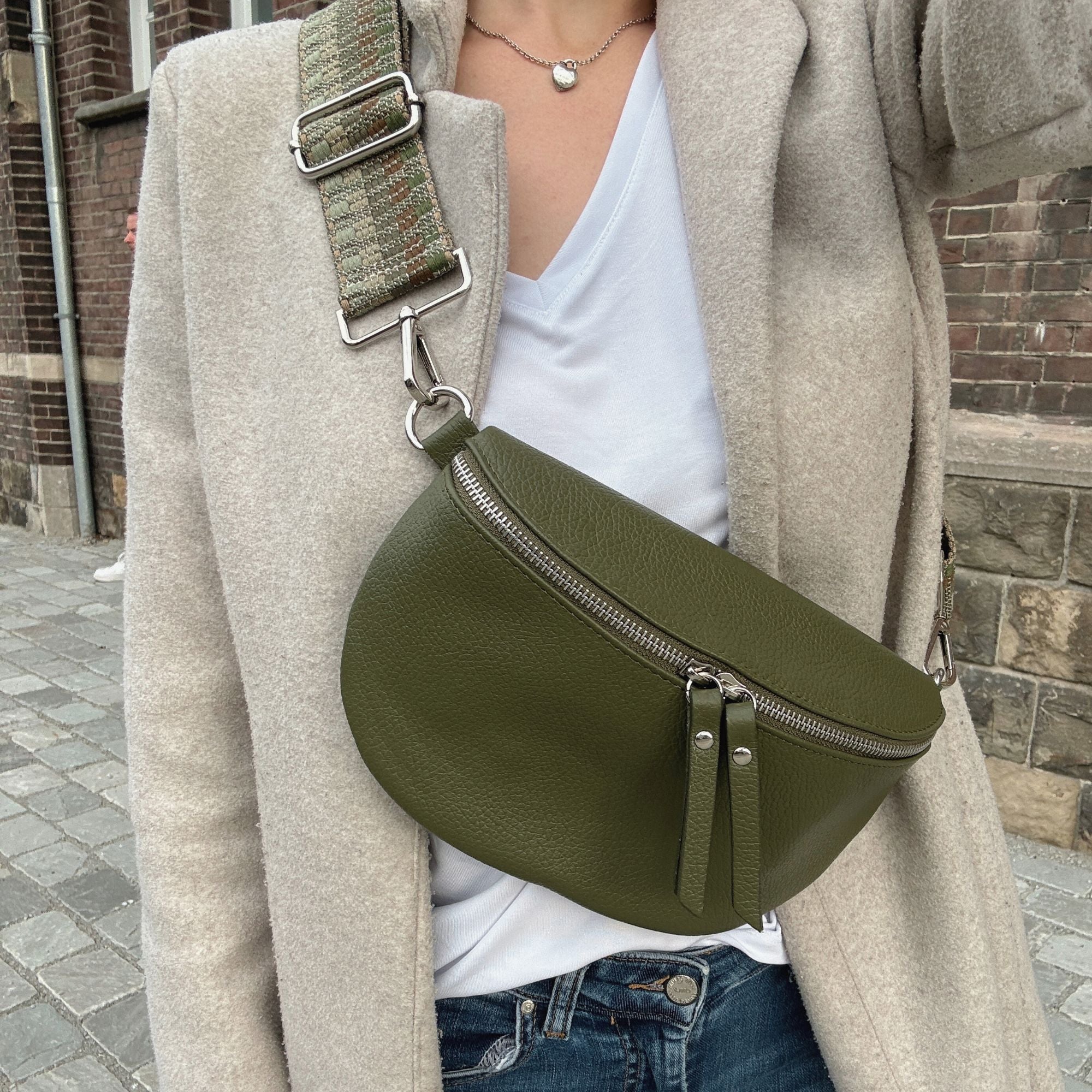 Selfie von Frau, welche beigen Mantel, weißes Shirt und die dunkelgrüne Leder Bauchtasche von MAONI trägt. Dazu kombiniert die Frau einen grün braunen Taschengurt.