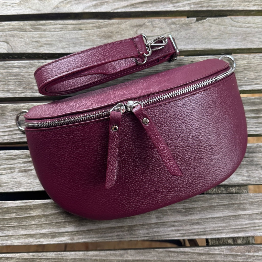Große burgunderrote Bauchtasche mit zwei Reißverschlüssen, auf Holztisch liegend – schmaler Lederwechselgurt diagonal darübergelegt