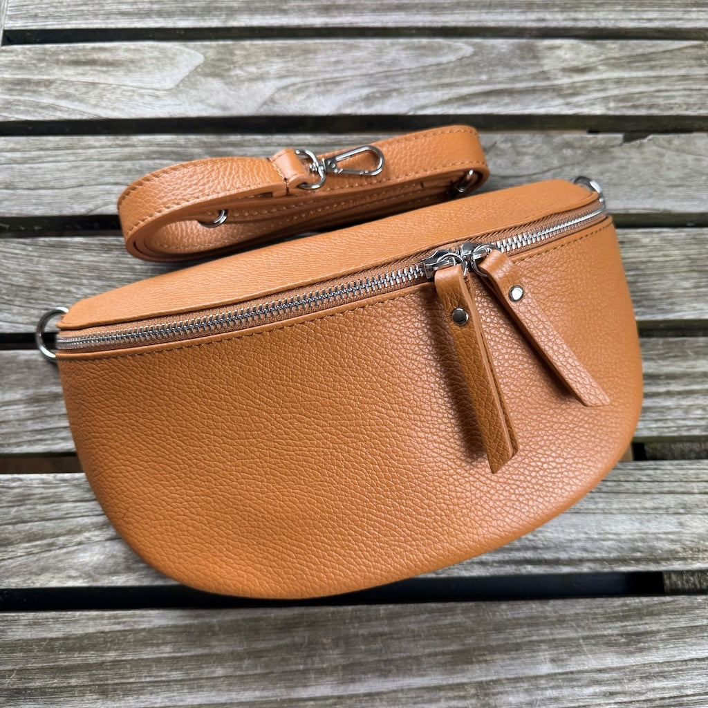 Große cognacfarbene Bauchtasche aus Leder, frontal auf Holztisch platziert – schmaler Ledergurt über der Tasche arrangiert