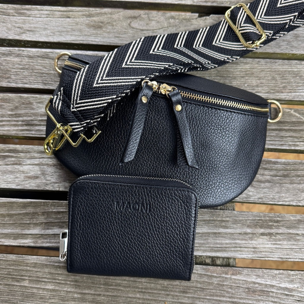 Schwarze Bauchtasche aus Leder auf Holztisch, darüber ein breiter gemusterter Gurt – unten im Bild eine schwarze Leder-Geldbörse mit geprägtem MAONI-Logo