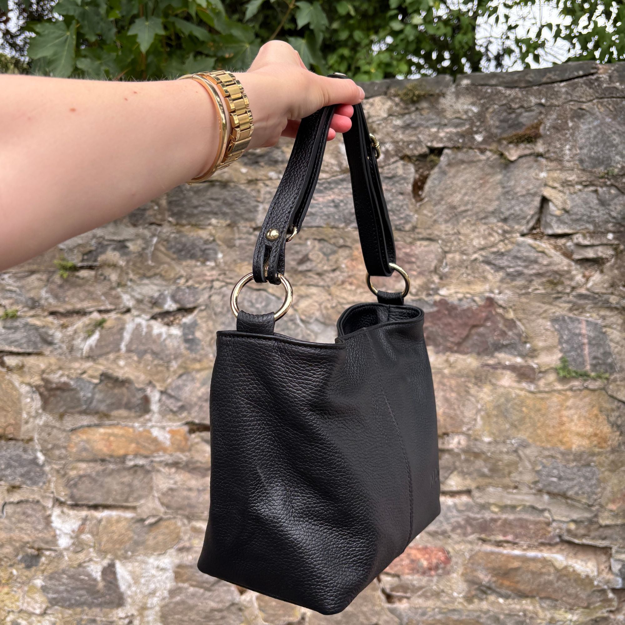 Schwarze Ledertasche Monica am kurzen Henkel vor einer Steinmauer