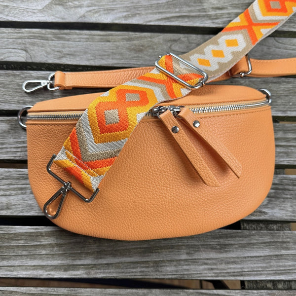 Flatlay auf Holztisch: Pfirsichfarbene Bauchtasche mit schmalem Ledergurt und breitem gemustertem Gurt in den Farben beige, taupe und orange – alle Teile übersichtlich arrangiert