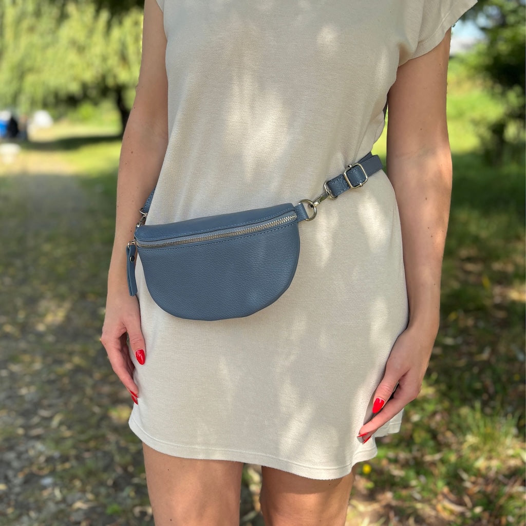 Blaue Bauchtasche von Frau um die Hüfte getragen. Frau trägt beiges Kleid und steht im Wald. 