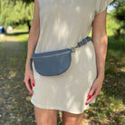 Blaue Bauchtasche von Frau um die Hüfte getragen. Frau trägt beiges Kleid und steht im Wald. 